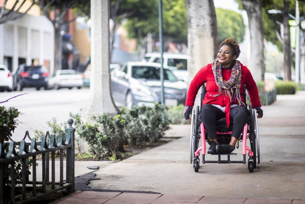 Una joven negra discapacitada con una silla de ruedas y un suéter de colores brillantes en una acera de la ciudad.